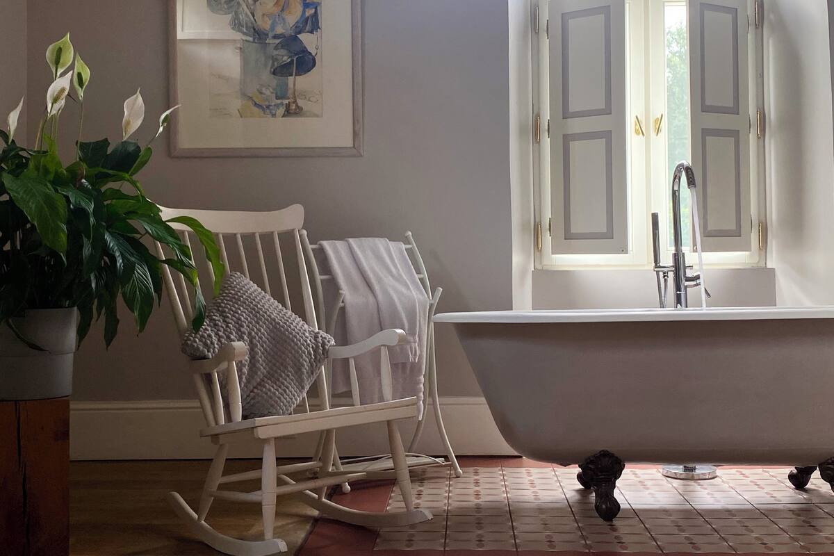 A bathroom with a white bathtub, a rocking chair, a potted plant, and a framed artwork on the wall.