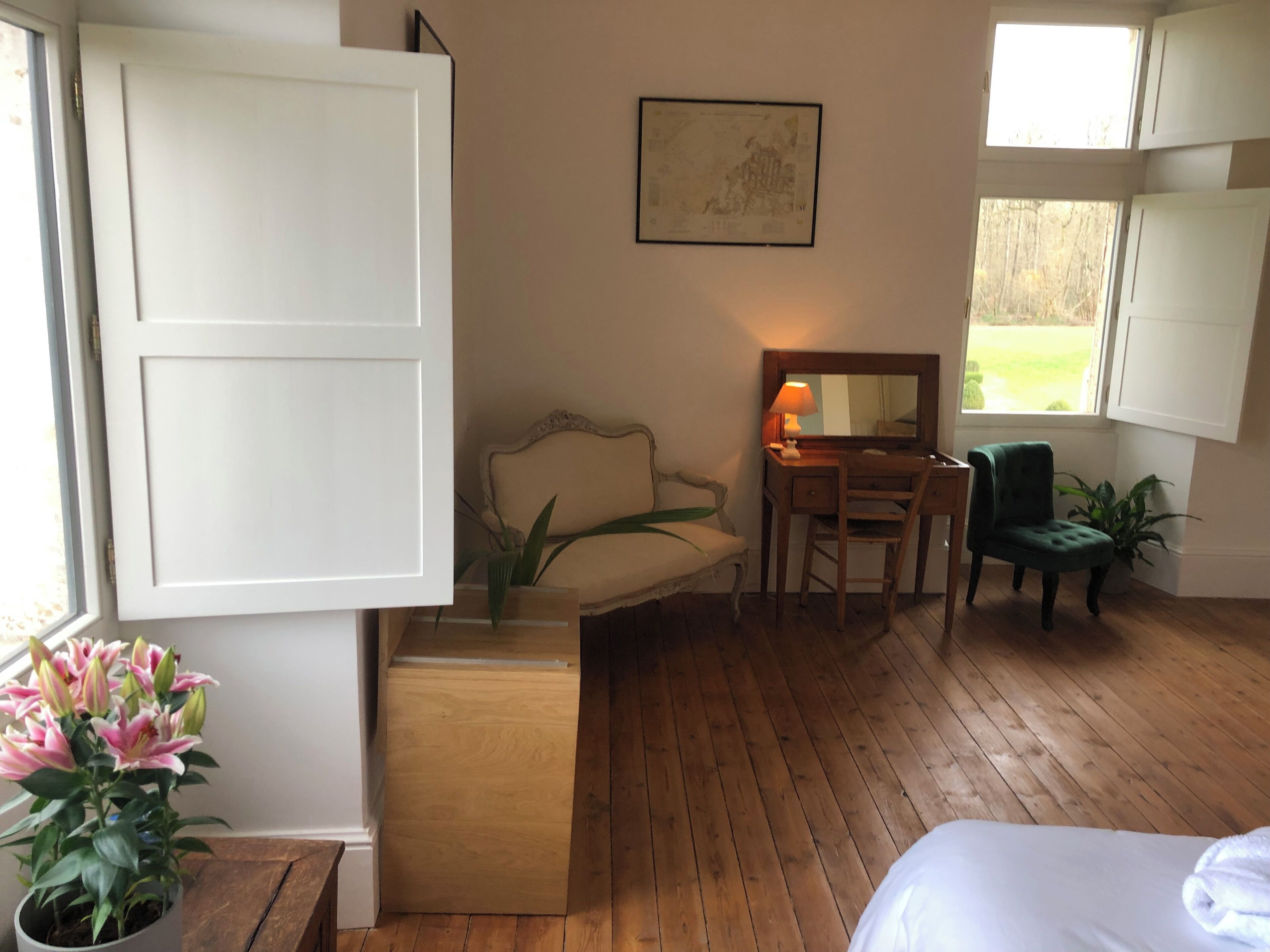 A room with a wooden floor, a bed, a desk with a chair, a green chair, a potted plant, and a window.