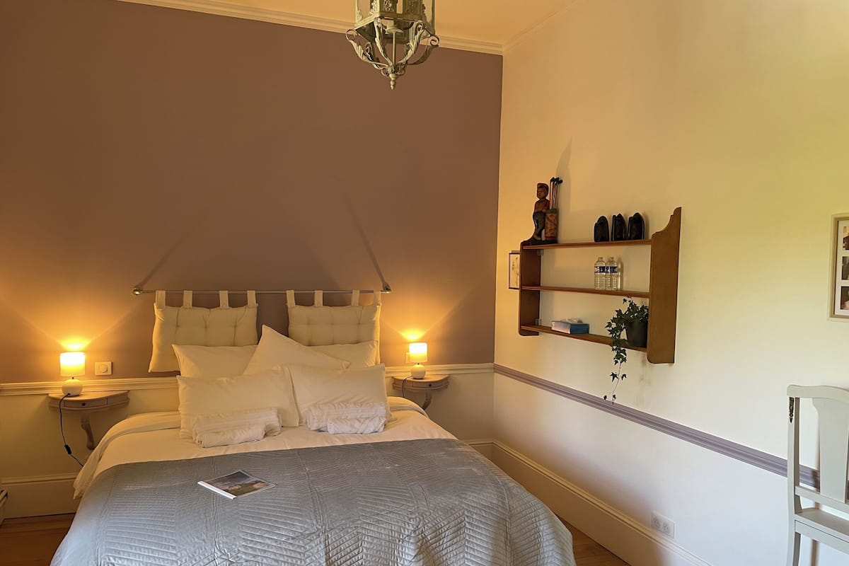 A bedroom with a large bed, two bedside lamps, a wall-mounted shelf with items, and a hanging light fixture.