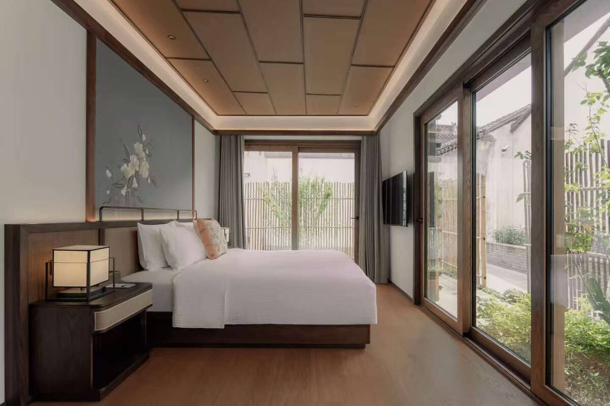 Une chambre d’hôtel moderne dotée d’un grand lit, d’une table de chevet avec une lampe, d’un téléviseur à écran plat et d’une grande fenêtre donnant sur l’extérieur.