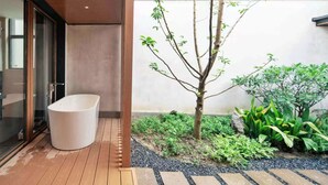 Une terrasse en bois agrémentée d’un jardinier blanc et d’un petit arbre, entourée de différentes plantes et bordée d’un chemin en pierre.