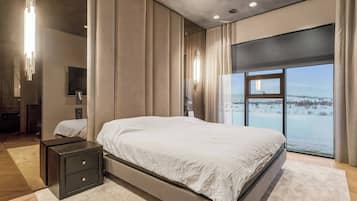 A modern bedroom with a large bed, a nightstand, a mirror, and a view of a snowy landscape through a large window.