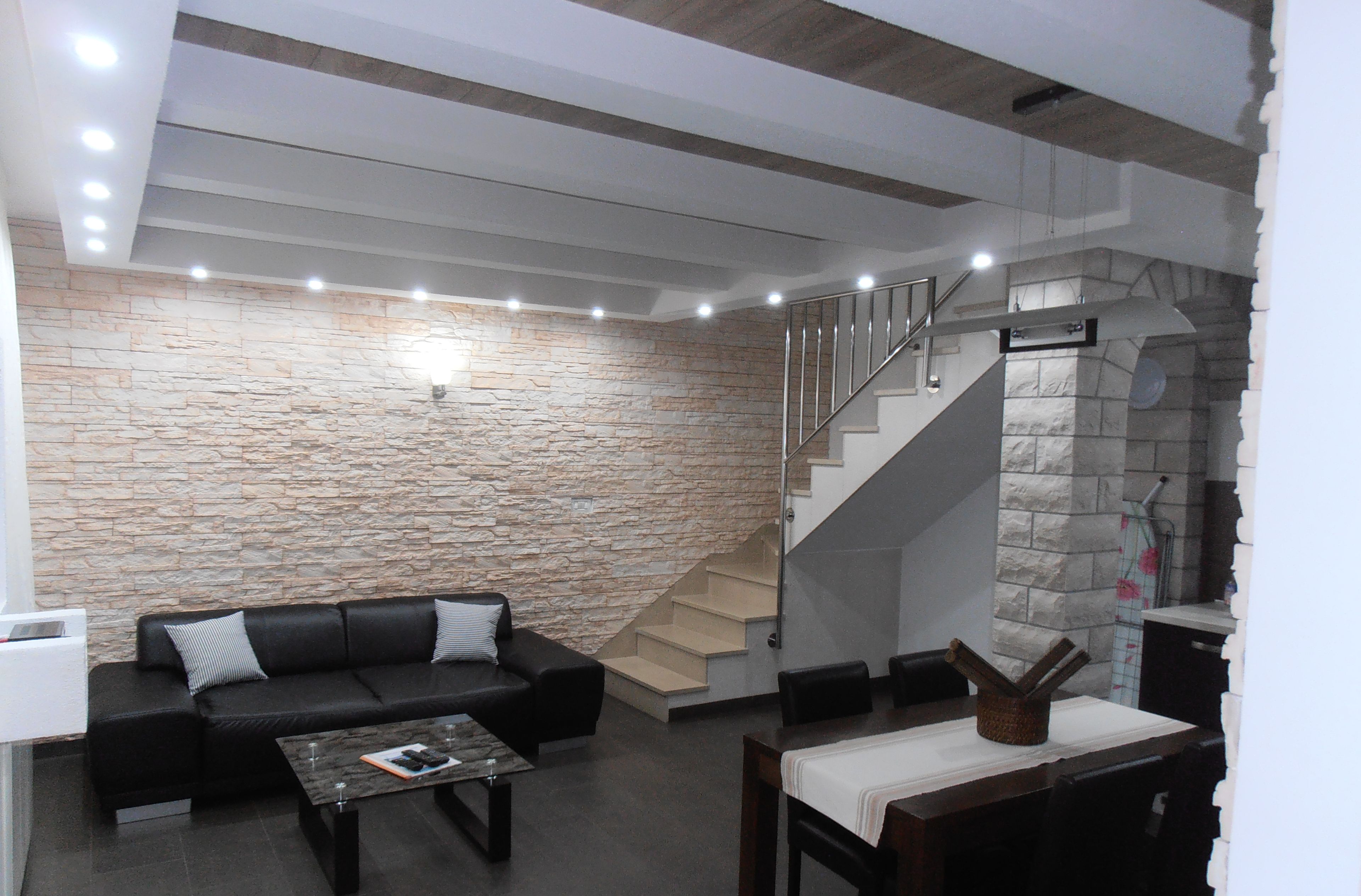 A modern living room with a stone wall, a black leather sofa, a coffee table, and a staircase leading to an upper level.