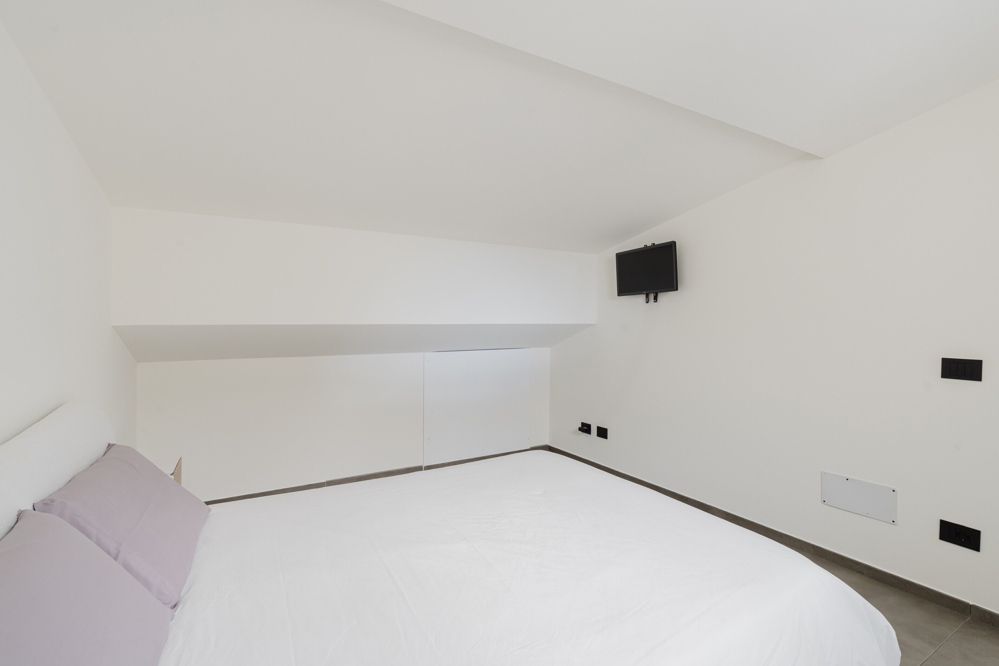 A minimalist bedroom with a bed, a television mounted on the wall, and a slanted ceiling.
