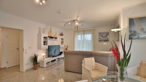 A modern living room with a sofa, two armchairs, a glass coffee table, a flat-screen TV, a ceiling fan, and decorative items including a ship model and a vase with flowers. - Ostsee Domizil, App. 1 (Timmendorfer Strand)