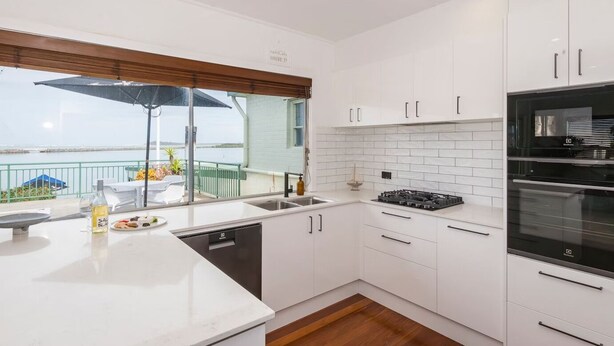 A modern kitchen with white cabinetry, a built-in oven, and a view of the sea. - The Happy Pelican Harrington Hotel (Harrington)