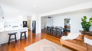 A modern kitchen with white cabinets, a central island with stools, a dining area with a wooden table and chairs, and a living room with a leather sofa and a large plant. - The Happy Pelican Harrington Hotel (Harrington)