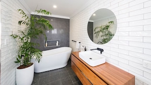 A modern bathroom with a freestanding bathtub, a wooden vanity, a round mirror, and a potted plant. - The Happy Pelican Harrington Hotel (Harrington)