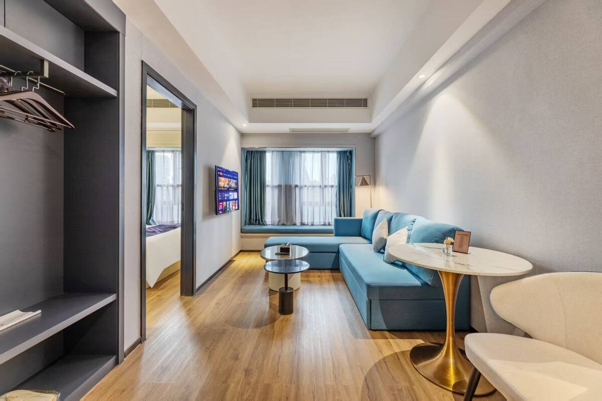 Ein modernes Hotelzimmer mit einem blauen Sofa, einem runden Couchtisch, einem kleinen Esstisch und einem an der Wand montierten Fernseher.