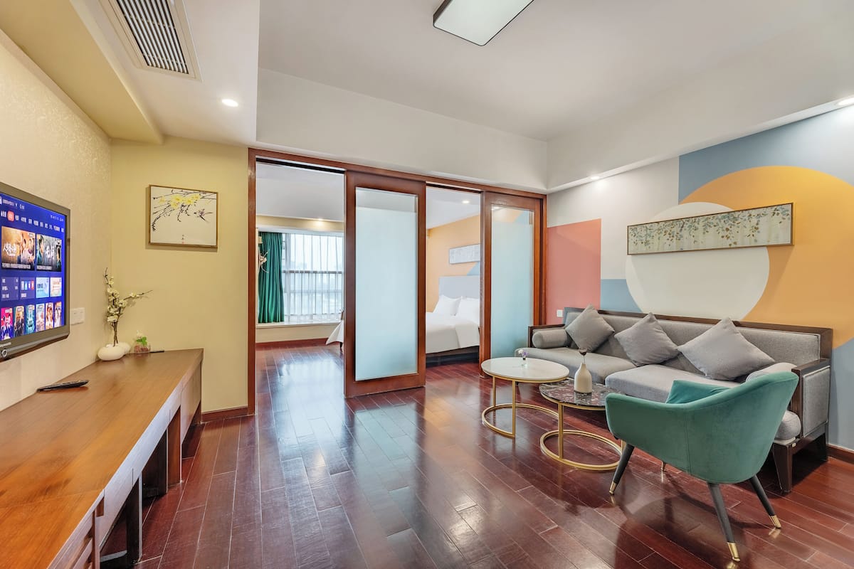 Ein modernes Hotelzimmer mit einem Holztisch, einer Couch und einem grünen Sessel. An der Wand hängt ein Fernseher und im Hintergrund steht ein Bett.