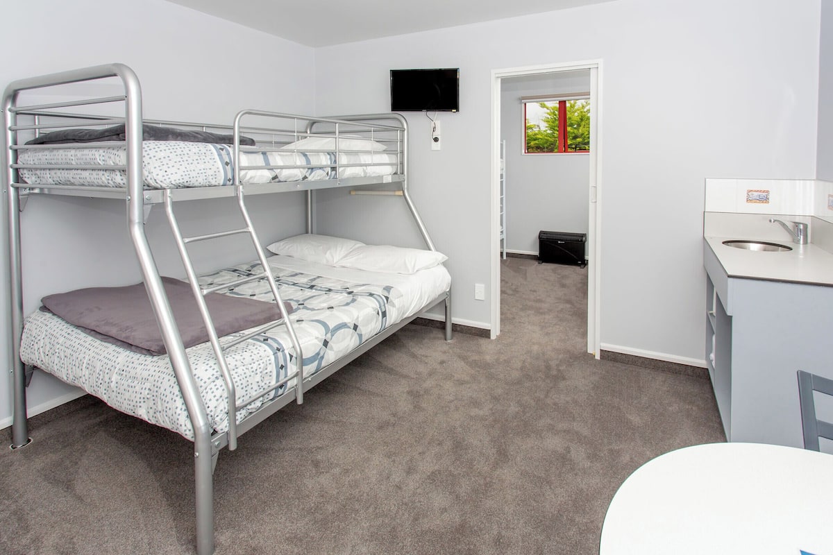 Una cama individual superpuesta con un televisor montado arriba, una kitchenette pequeña con un lavamanos y una puerta que da a un baño.