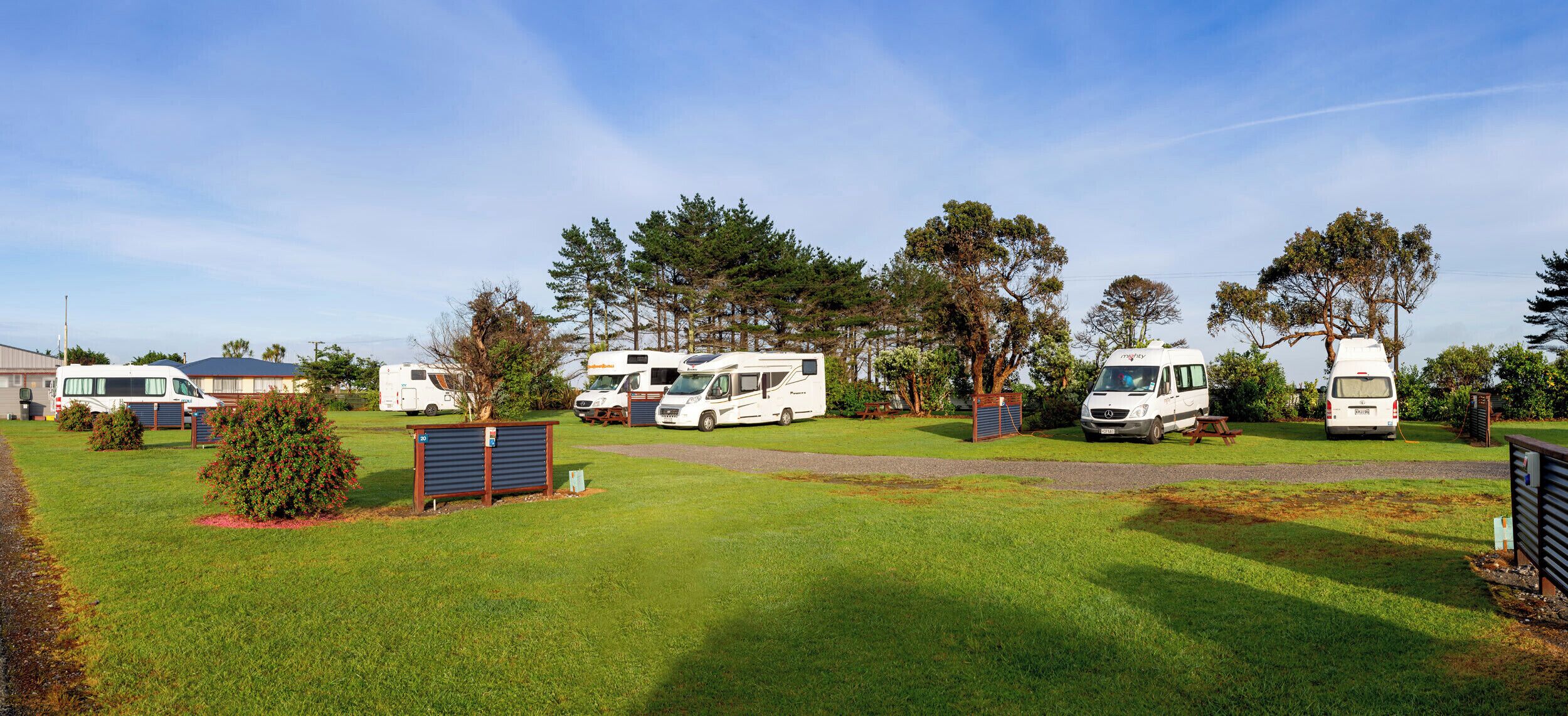 Un terrain de camping où des camping-cars sont stationnés sur une pelouse, entourés d’arbres et sous un ciel dégagé.