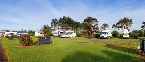 Un terrain de camping où des camping-cars sont stationnés sur une pelouse, entourés d’arbres et sous un ciel dégagé.
