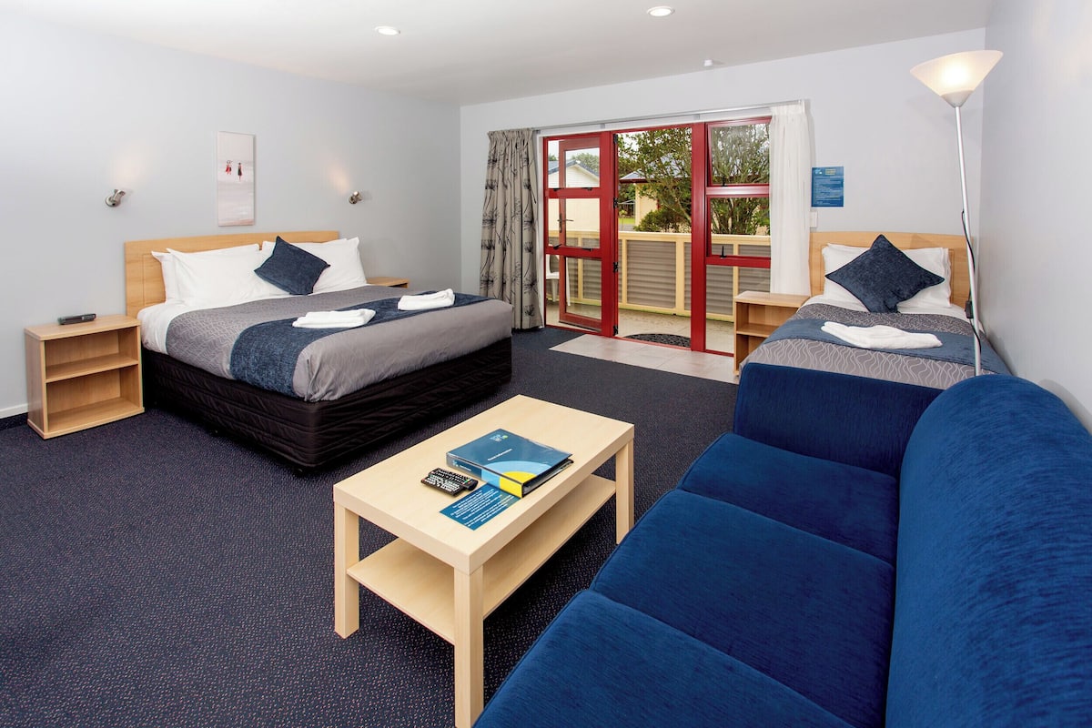 Habitación de hotel con dos camas, un sofá, una mesita y un ventanal corredero al exterior.