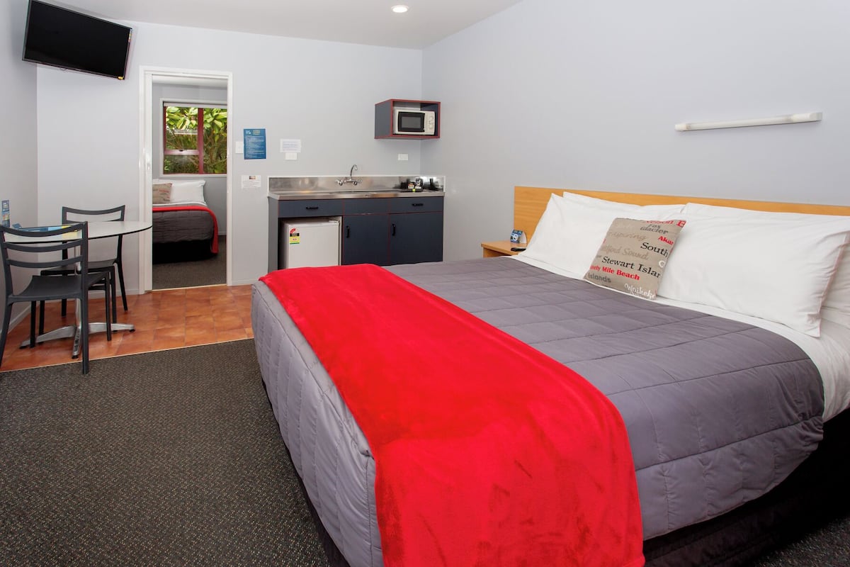 Habitación de hotel con cama, escritorio con silla, kitchenette pequeño y televisor montado en la pared.