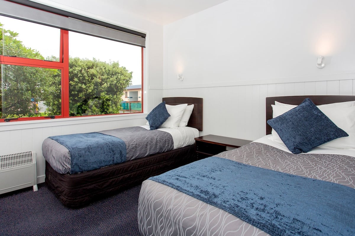 Habitación de hotel con dos camas individuales, un ventanal grande con marco rojo y vistas a zonas verdes.