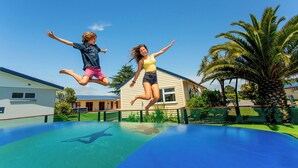 Two people jumping on a trampoline in a backyard with a house and palm trees. - Carters Beach Top 10 Holiday Park (Westport)