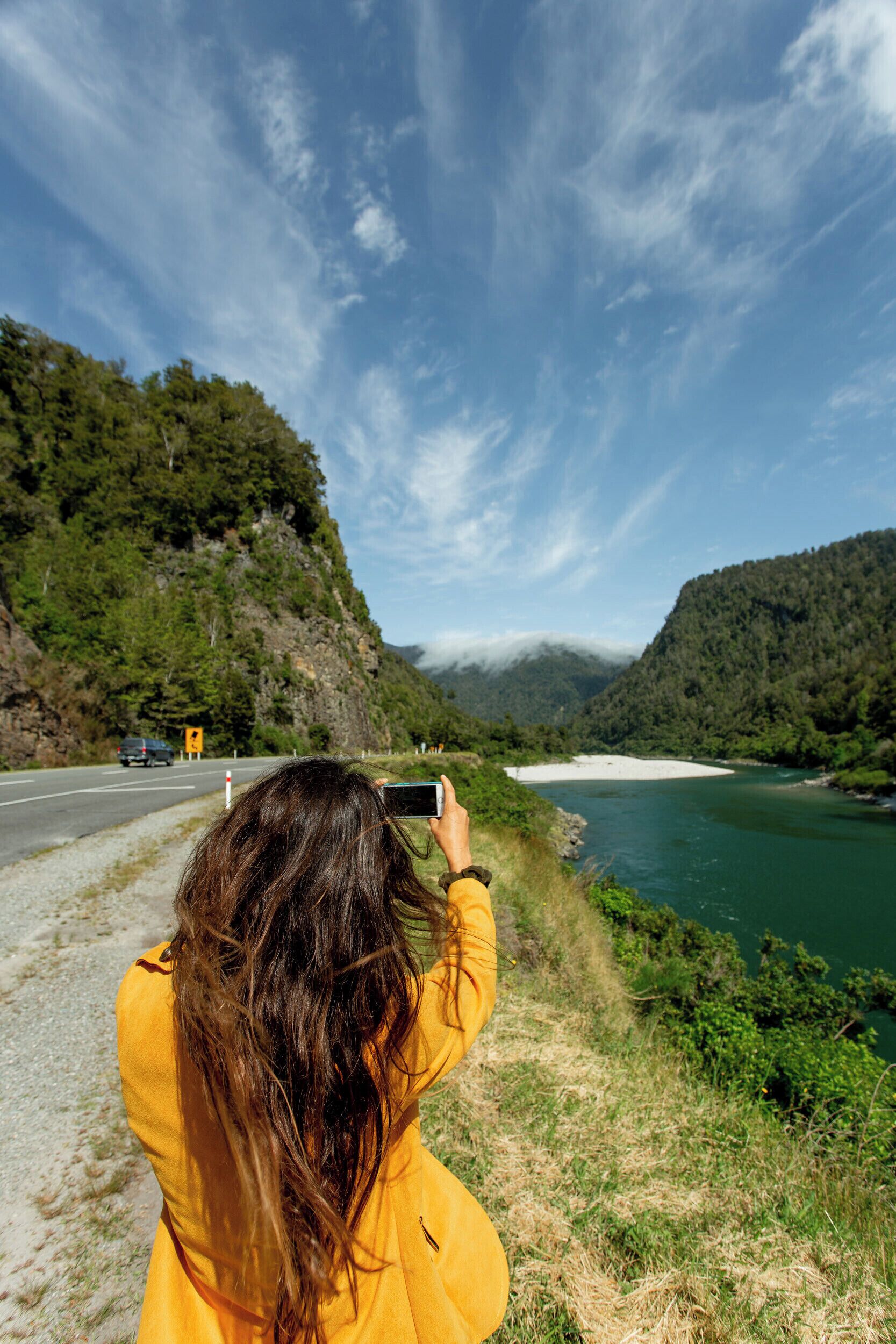 A person taking a photo of a river with mountains in the background.