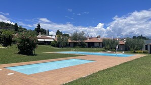 A pool area with two rectangular pools, surrounded by a wooden deck, and a grassy lawn with a building in the background. - Harmonia Cottage by Bookinggardalake (Soiano del Lago)