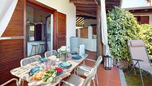 An outdoor dining area with a table set for two, surrounded by white wicker chairs, a wooden pergola overhead, and a lush green hedge. - Harmonia Cottage by Bookinggardalake (Soiano del Lago)