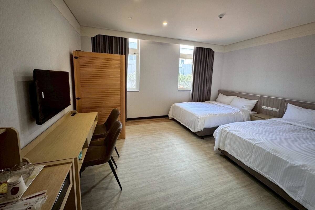 A hotel room with a desk, two beds, a TV, and a window.
