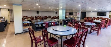 A spacious dining area with multiple round tables and red chairs, a buffet station, and a kitchen area.