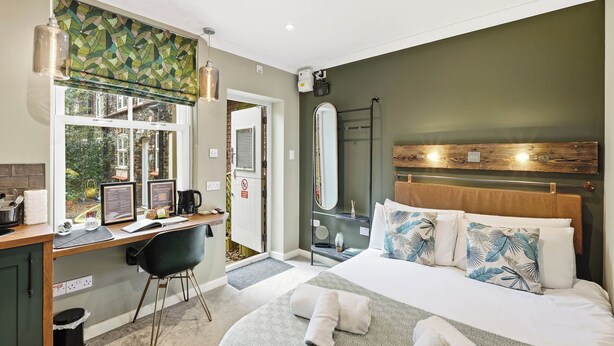 Una habitación de hotel moderna con una cama grande, un escritorio con computadora, una ventana con vistas a zonas verdes y un espacio con ducha.