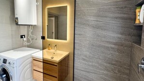 A modern bathroom with a built-in washing machine, a vanity with a mirror, and a shower area with a dark tiled wall.
