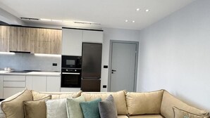 A modern living room with a beige sofa and cushions, a kitchen with white cabinets and stainless steel appliances, and a door to the right.
