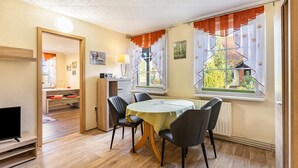 A dining area with a round table, chairs, a television, and a view of a garden through the window. - Holiday Home Near Forest and Lake (Templin)
