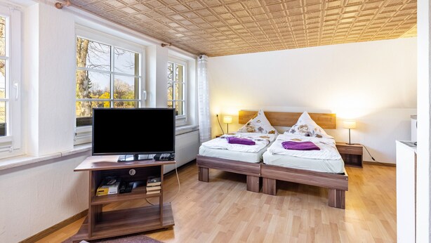A hotel room with a TV on a wooden stand, two beds with pillows, a bedside lamp, and a window with a view of trees. - Apartment 'Für Monteure' with Wi-Fi (Templin)