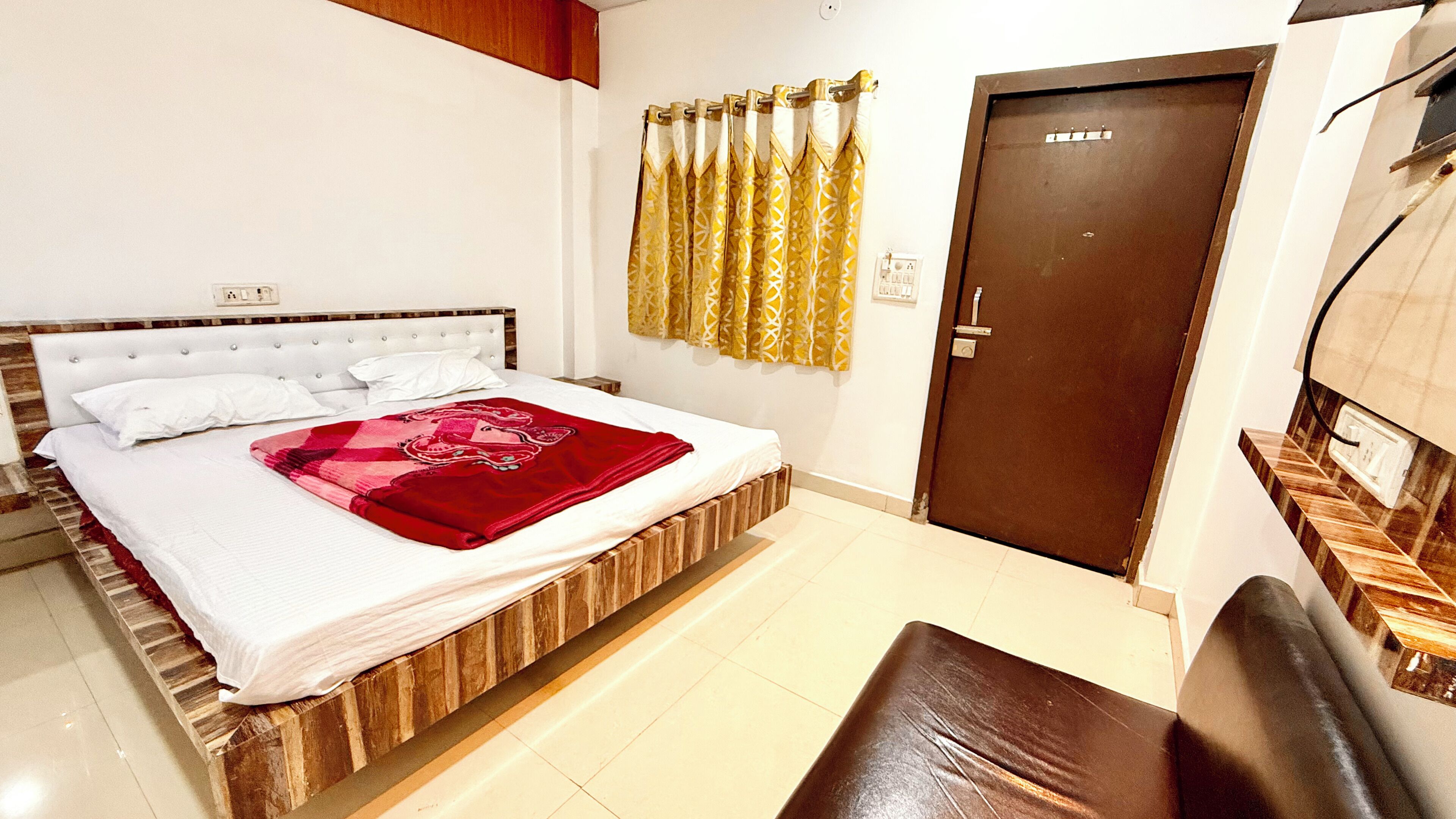 A bedroom with a bed, a wooden headboard, a red blanket, a brown door, and a brown bench.