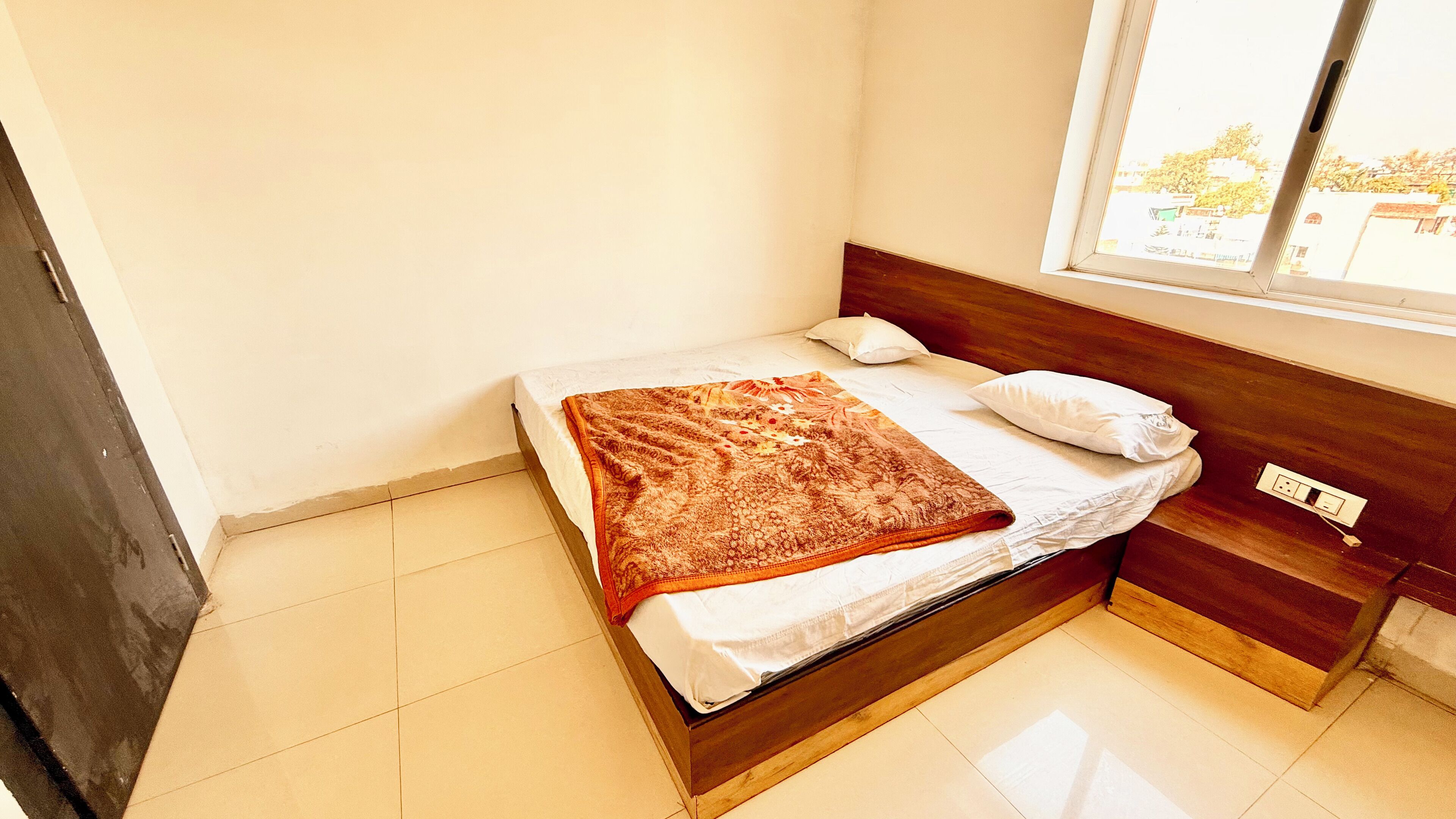 Un petit lit bien fait, avec une couverture à motifs, une tête de lit en bois et une fenêtre donnant sur les bâtiments environnants.