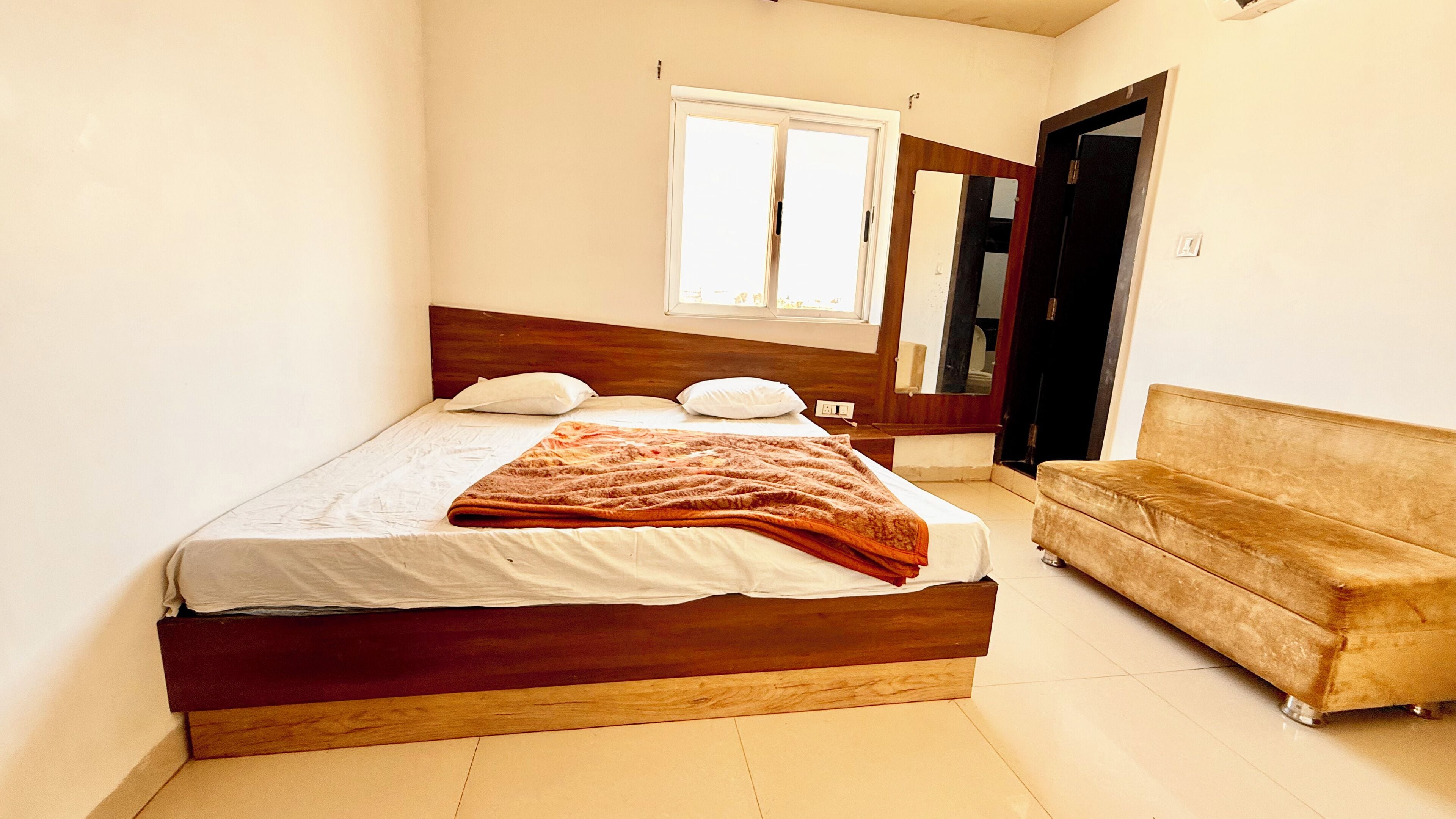 Un dormitorio con una cama individual, cabecera de madera, una mesita de noche, un sofá marrón, una ventana y un espejo en la pared.