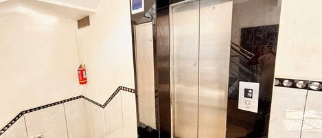 An elevator with a fire extinguisher mounted on the wall, a control panel, and a tiled floor with a black border.