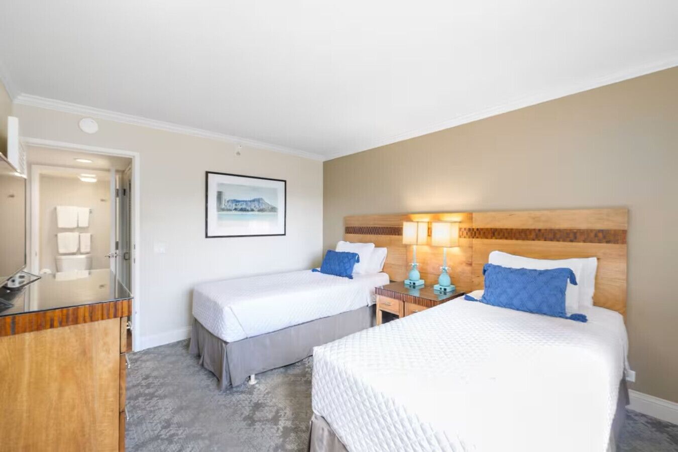 A hotel room with two beds, a wooden headboard, a bedside table with lamps, and a framed picture on the wall.