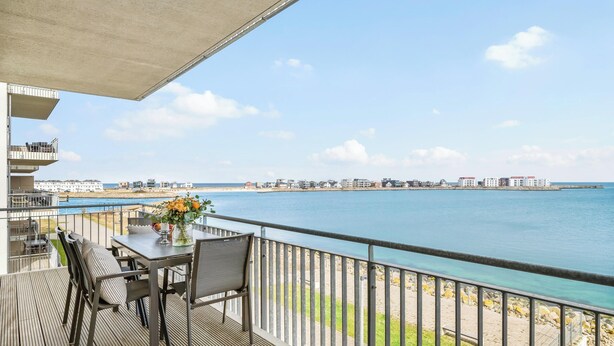 A balcony with a table and chairs, overlooking a coastal area with buildings and a clear sky. - Portus - Sea view, balcony, barbecue, walk-in shower, fireplace (Olpenitz)