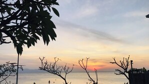 Une scène de plage où l’on voit des personnes assises et se promenant le long du rivage, des arbres et un coucher de soleil sur l’eau.