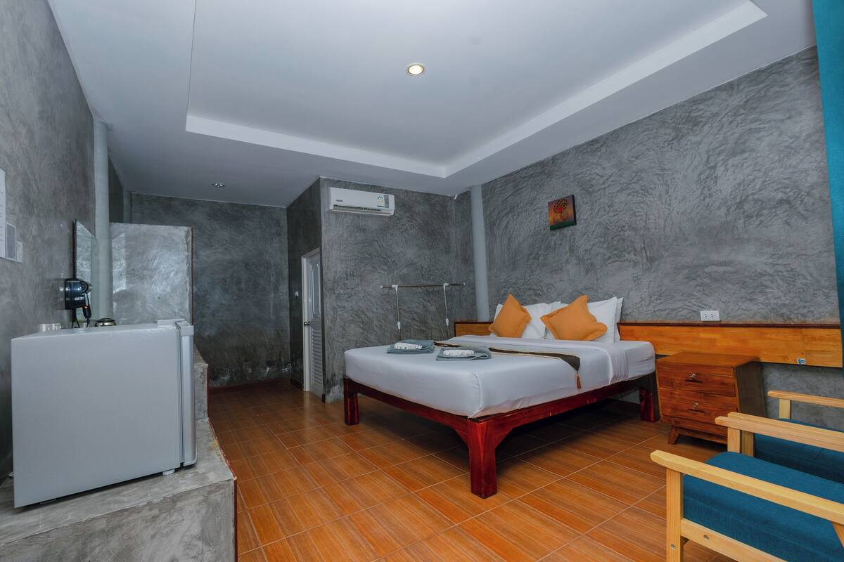 Quarto de hotel com cama, mesa de cabeceira de madeira, uma cadeira, uma mesinha e um frigorífico.