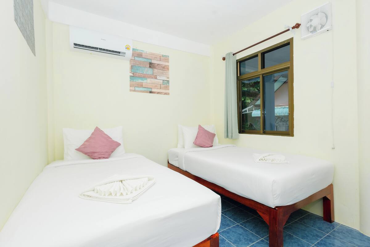 Quarto de hotel com duas camas de solteiro, cada uma com roupa de cama branca e almofadas cor-de-rosa. Há uma janela com vista para o exterior e um aparelho de ar condicionado fixado na parede.