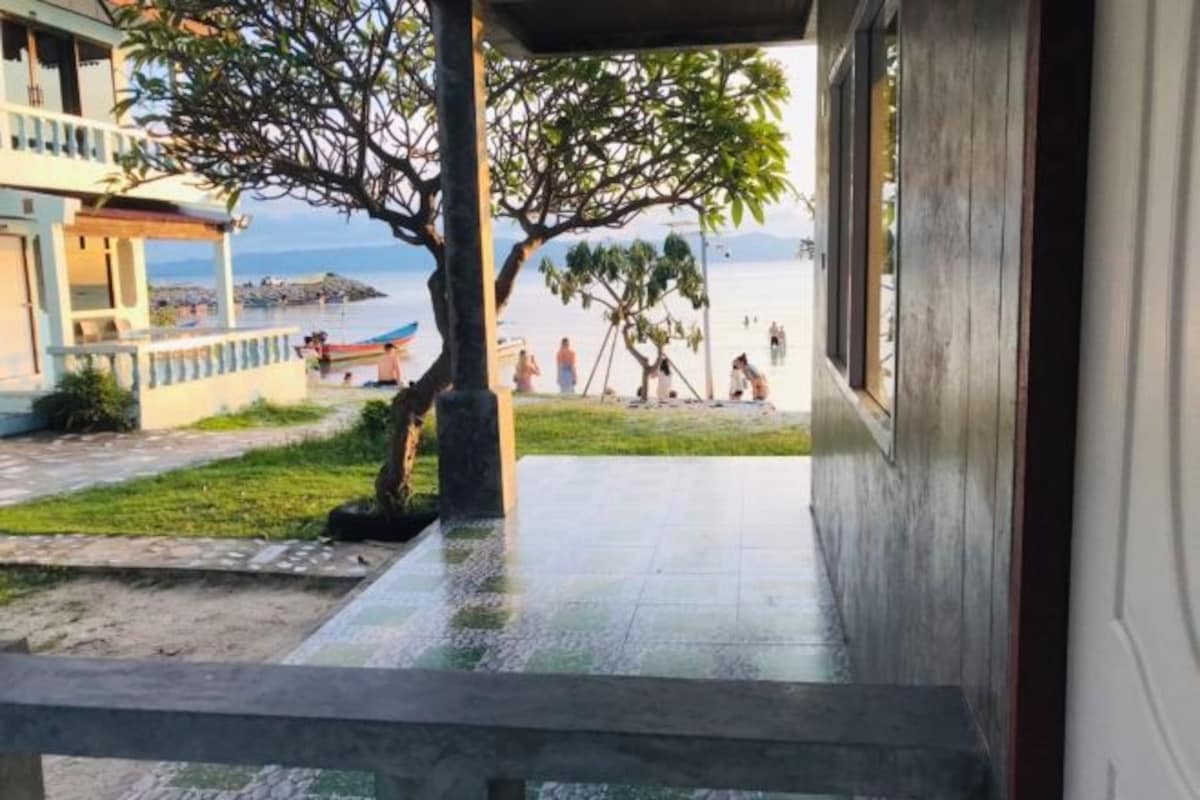 Vista desde un corredor techado hacia una playa, con un árbol y un edificio al frente.
