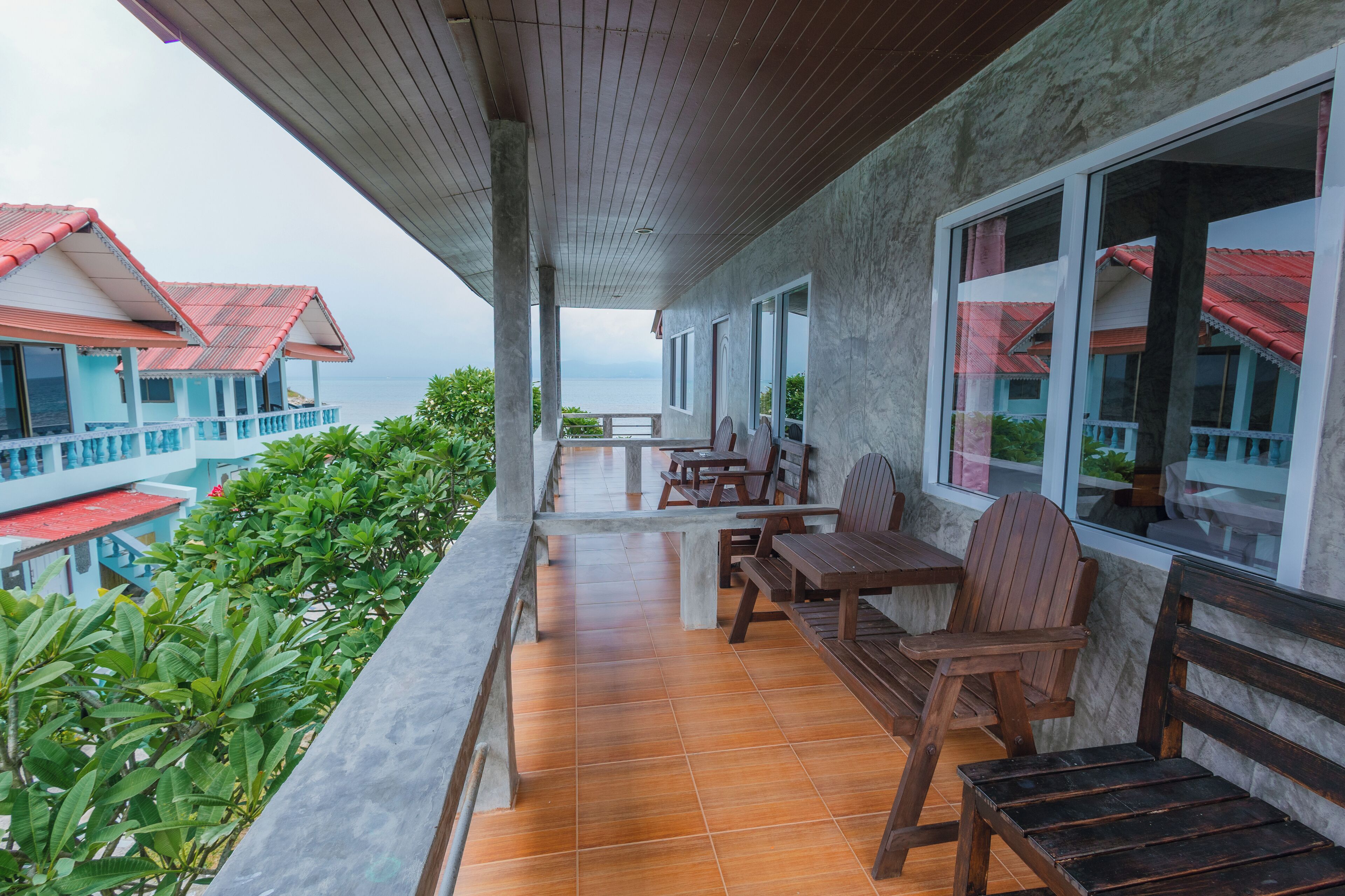 A covered outdoor area with wooden furniture, a view of the sea, and buildings with red roofs in the background.