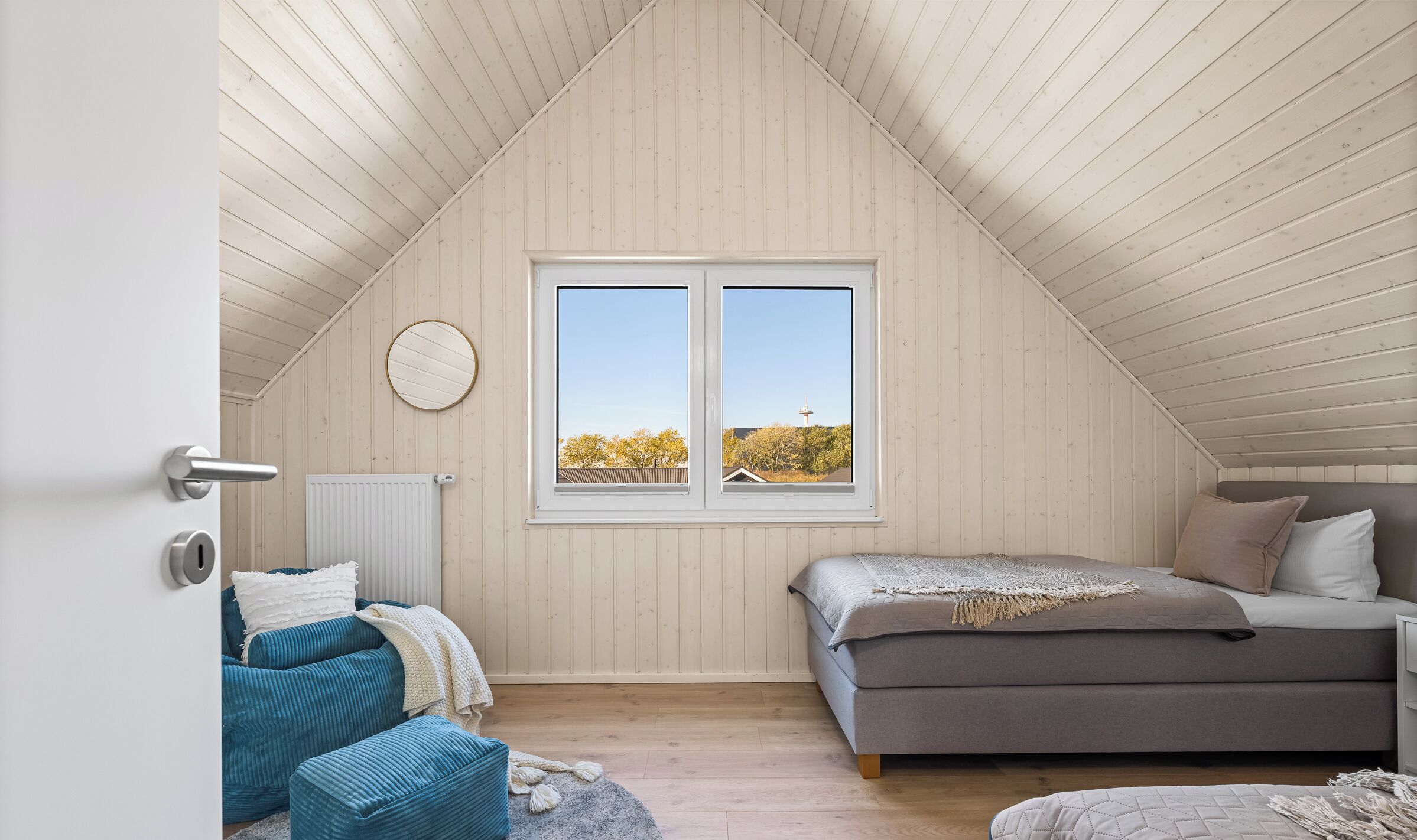 Um quarto com teto inclinado, uma janela grande com vista para árvores e uma torre, uma cama com almofadas, uma cadeira azul e um radiador.