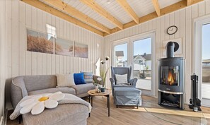 A cozy living room with a sofa, armchair, and a glass coffee table. A modern fireplace is built into the wall, and there are large windows offering a view of the outside.
