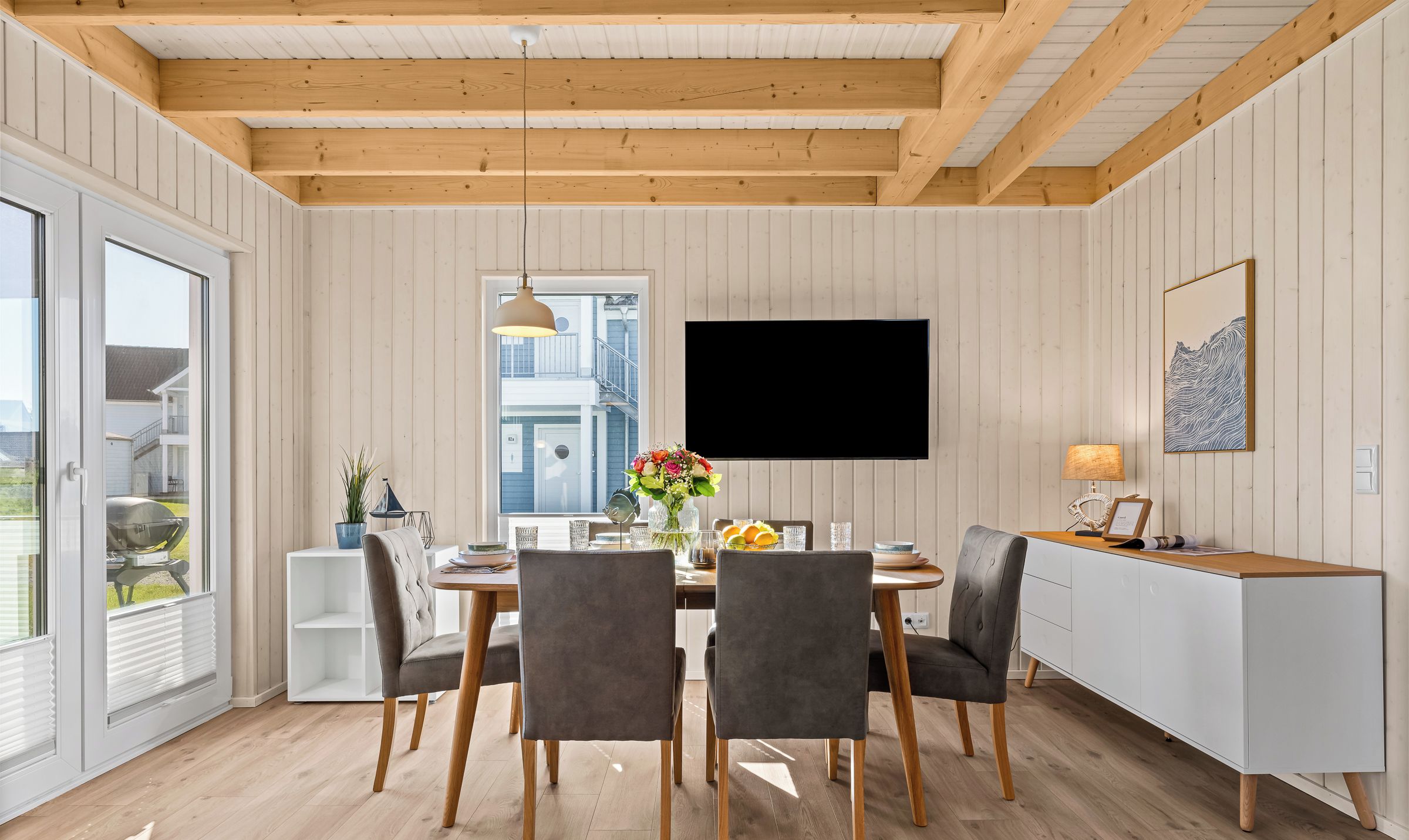 A dining area with a wooden ceiling, white paneled walls, a large flat-screen TV, and a modern white cabinet.