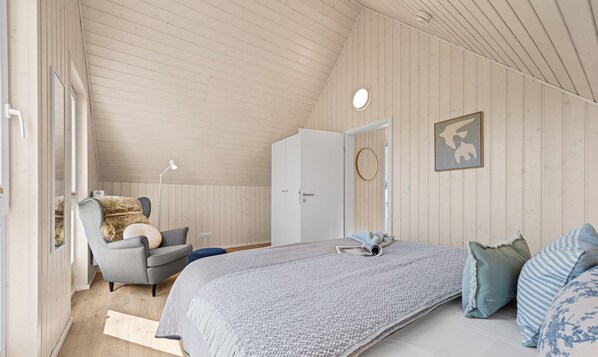 Ein Schlafzimmer mit schrägem Dach, einem Bett mit grauer Decke, einem Sessel mit Fellüberwurf und einer Tür mit runder Spiegelung.