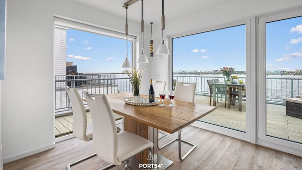 A modern dining area with a wooden table, white chairs, and pendant lights. It features large windows with a view of the water and cityscape. - Morgenlicht - Maisonette Apartment, Terrace, Infrared Sauna, Whirlpool, Sea View (Olpenitz)