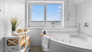 Un baño moderno con bañera, una ventana con vistas al mar y un estante con artículos de aseo.