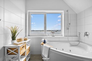 Une salle de bain moderne avec une baignoire, une fenêtre donnant sur la mer et une petite étagère contenant des articles de toilette.