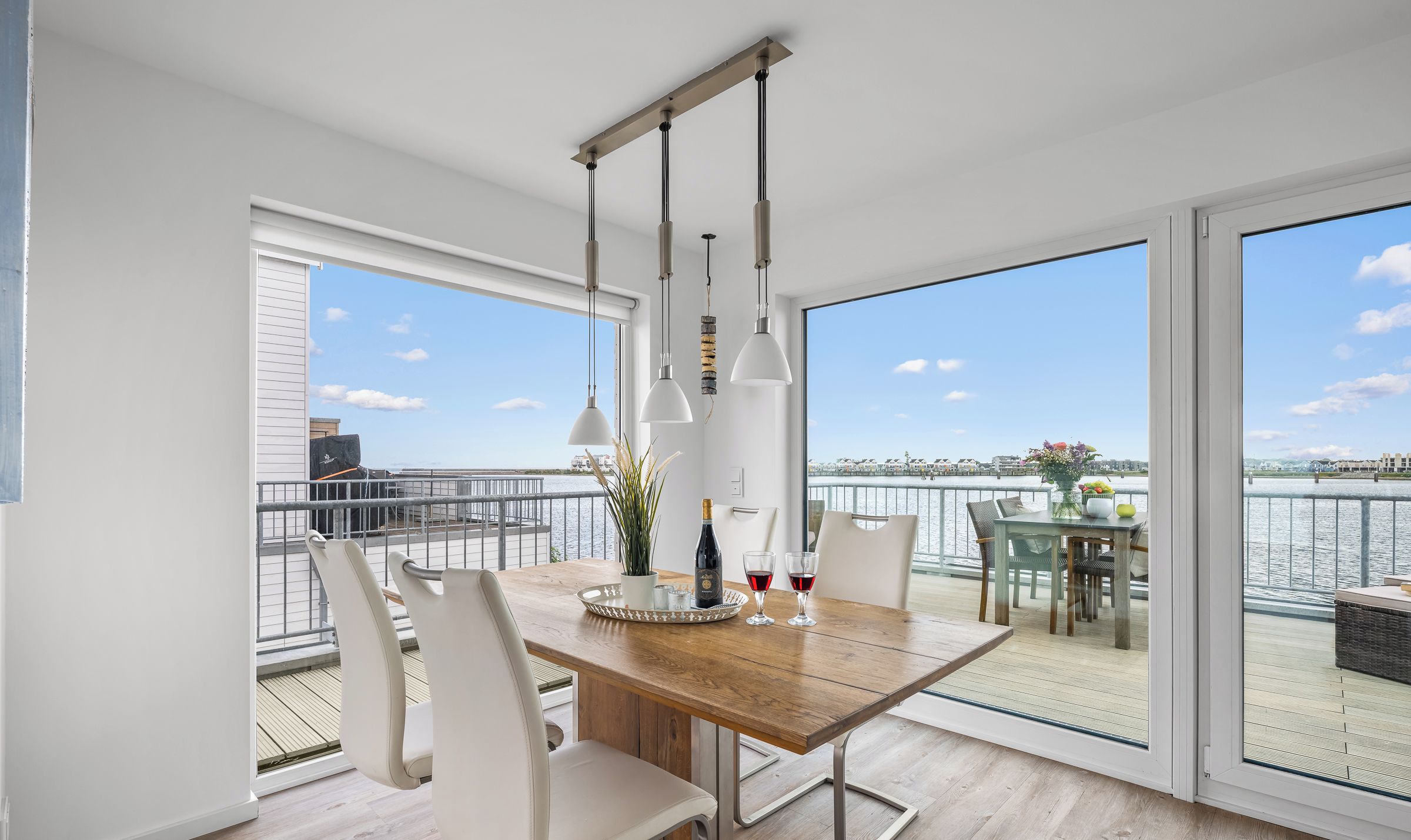 Un espace de repas moderne avec une table en bois, des chaises blanches et des luminaires suspendus. Il est doté de grandes fenêtres offrant une vue sur l’eau et le paysage urbain.